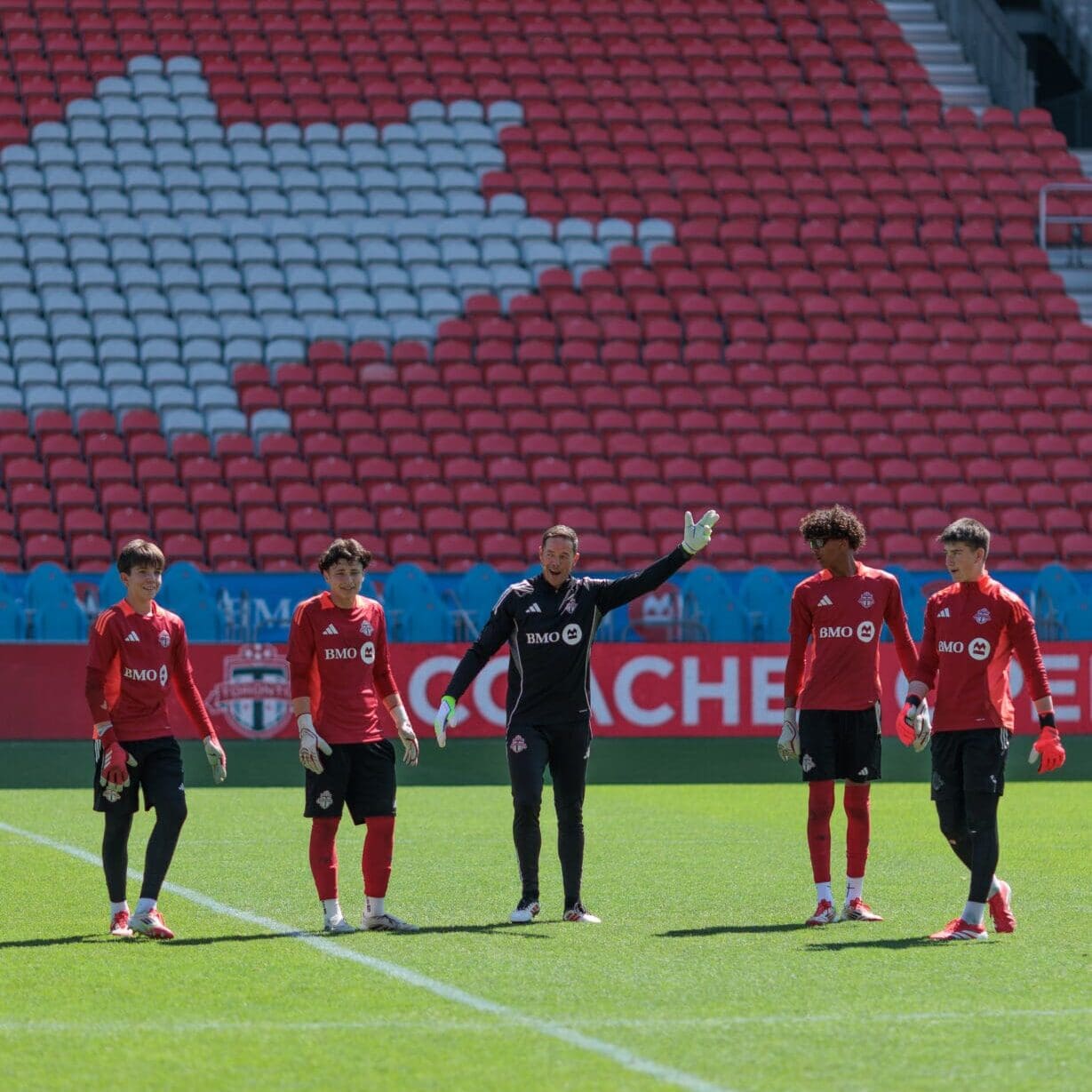 Soccer coach in all black stands in between four young soccer players wearing red jerseys and black shorts on a soccer field with stadium seating in the background.